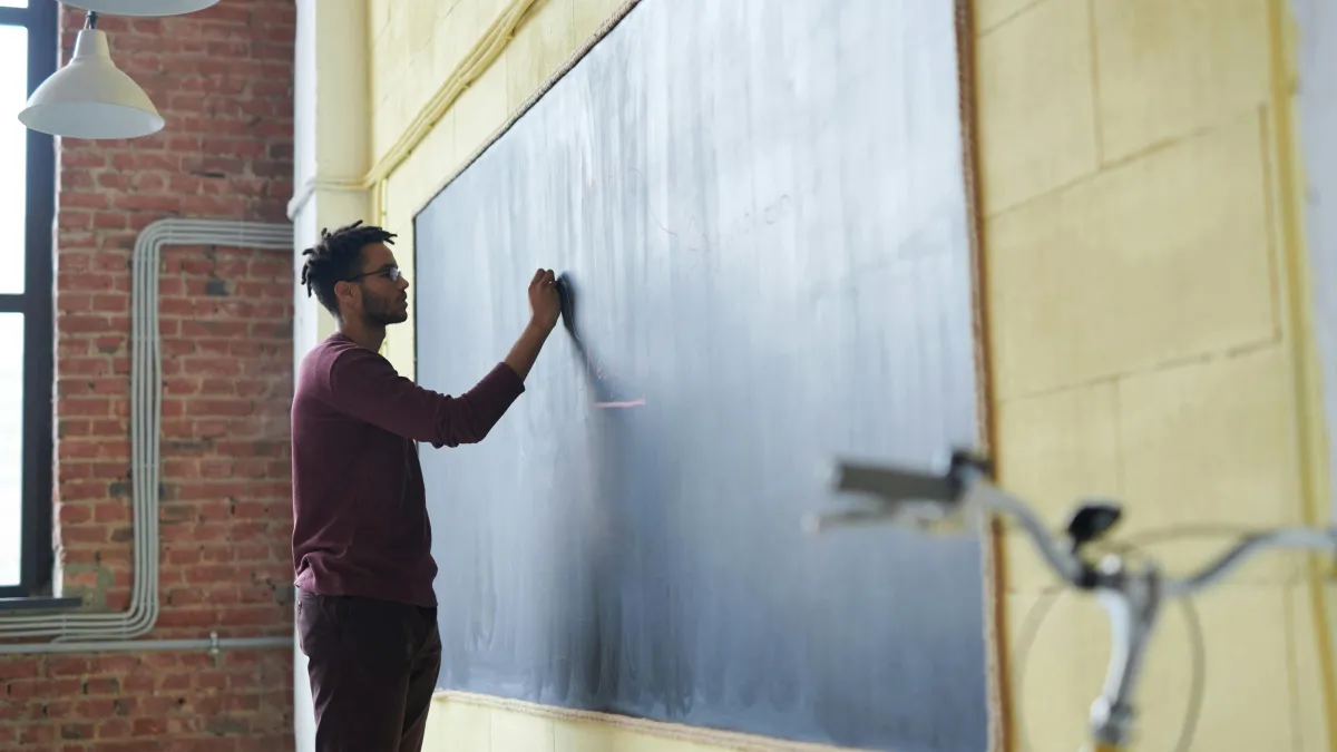 A man writes with chalk on a blackboard at the front of a classroom