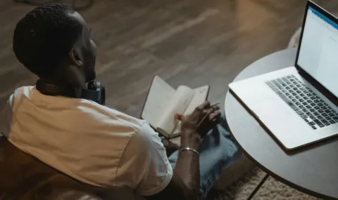 Thumbnail photo of man sitting with his computer open on a table and looking down at his notebook.